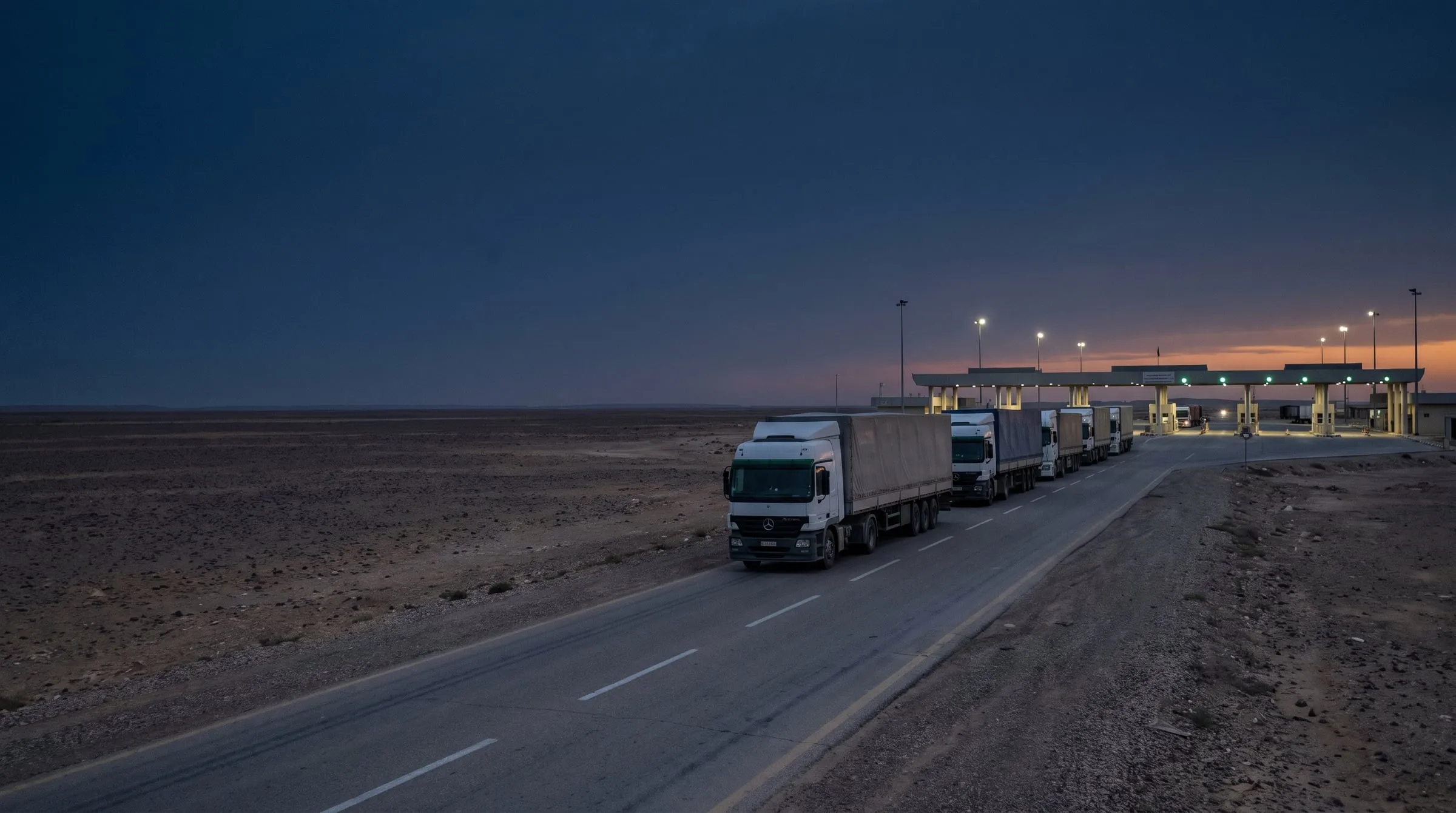Freight trucks on a desert highway approaching a land border crossing.