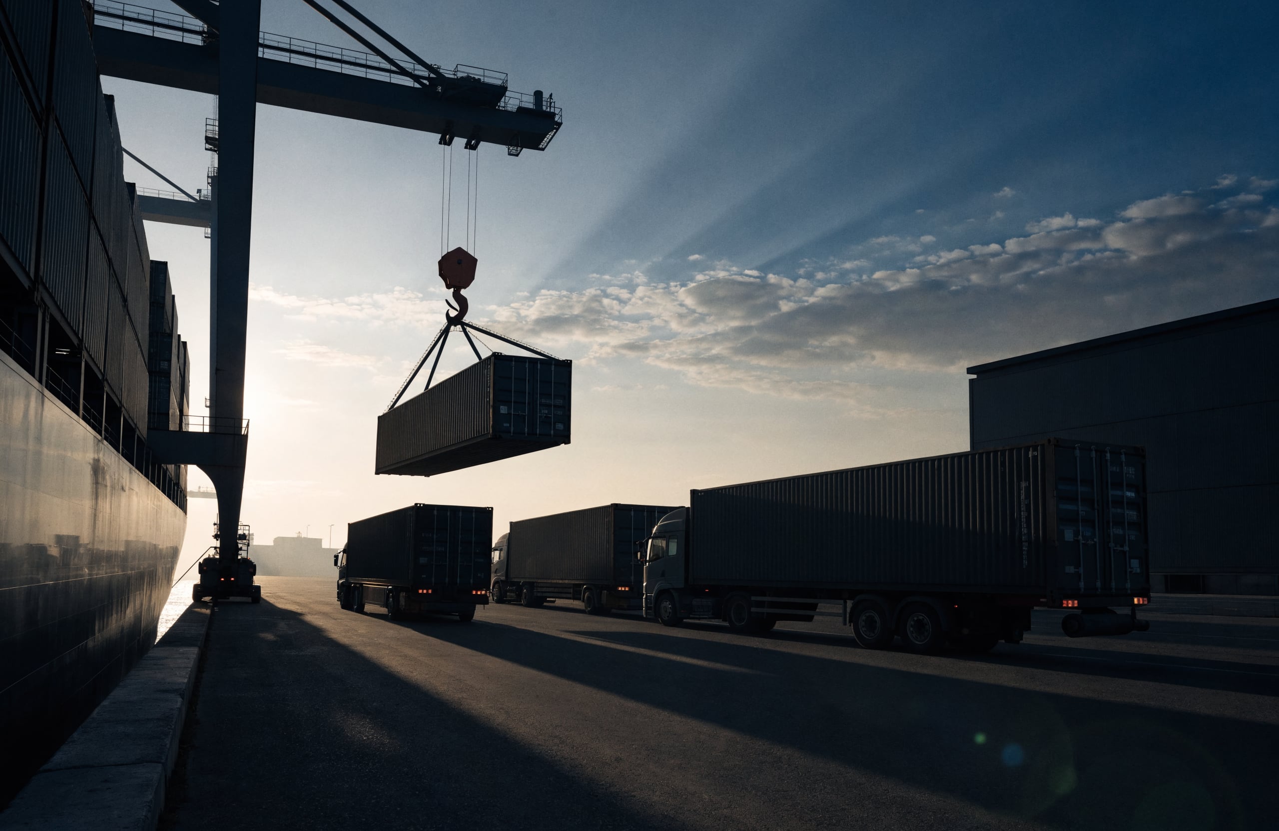 A container being lifted beside trucks at a port terminal.